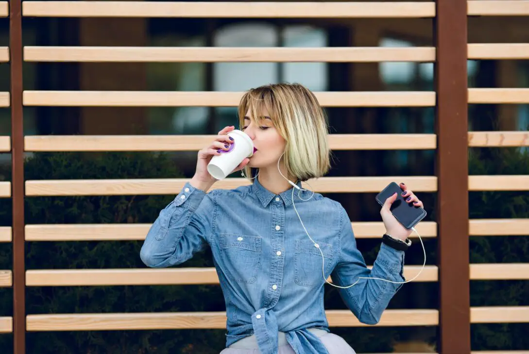A portrait of a blonde girl drinking a cup of coffee and listening to music on a smartphone with