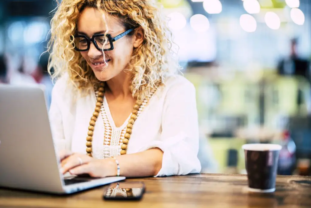 Happy woman working on laptop at coffee shop. Woman in eyeglasses and beads necklace