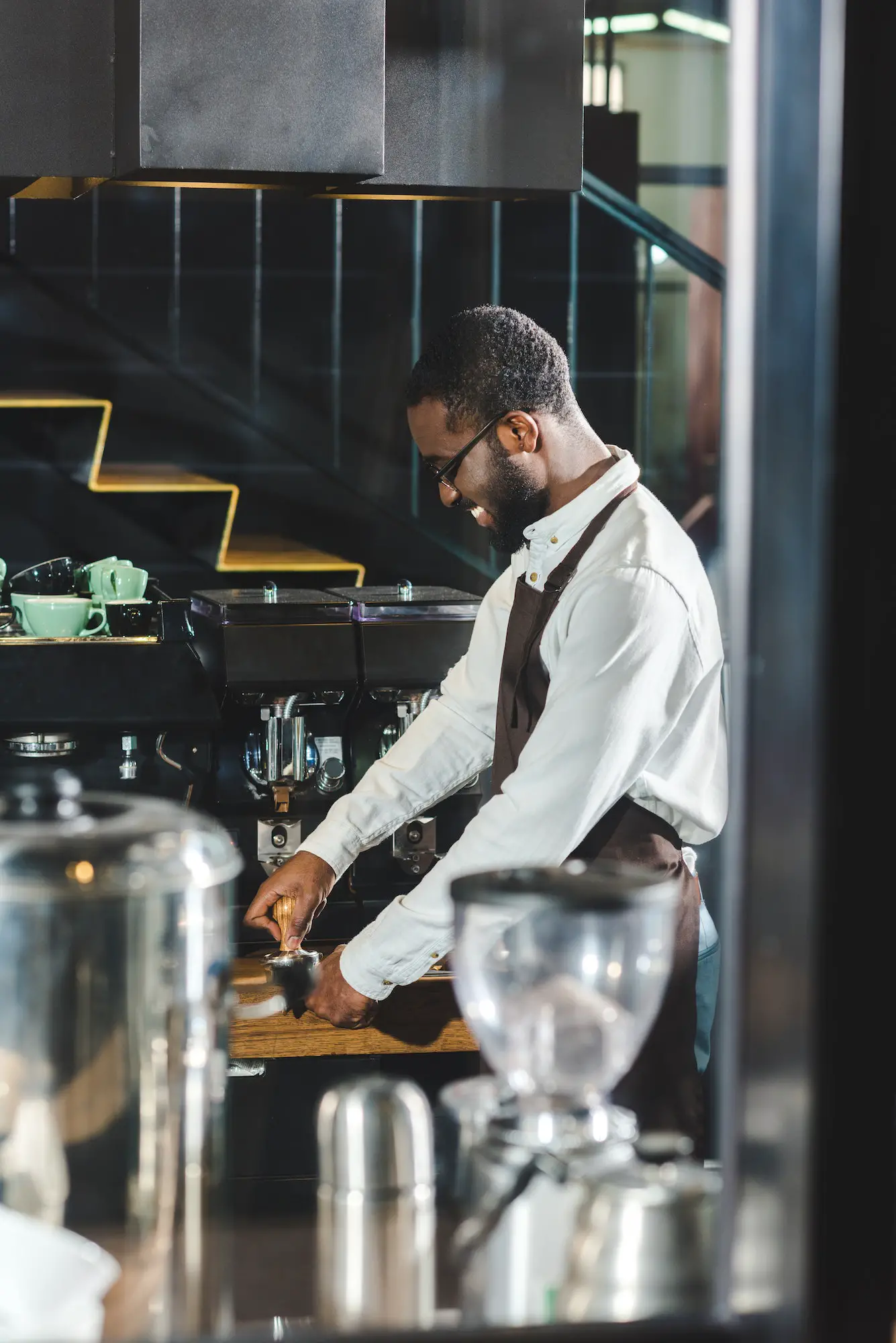 side view of smiling young african american barista in eyeglasses making coffee at coffee machine