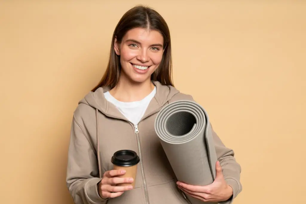 Woman fitness instructor posing after workout with rolled mat and plastic cup of coffee