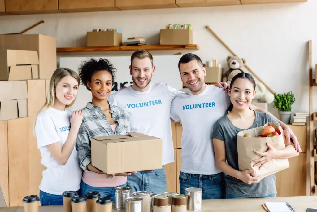 happy multicultural women holding cardboard box and paper bag with apples while standing near