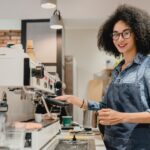 Smiling african young female barista making coffee using coffee machine in cafeteria